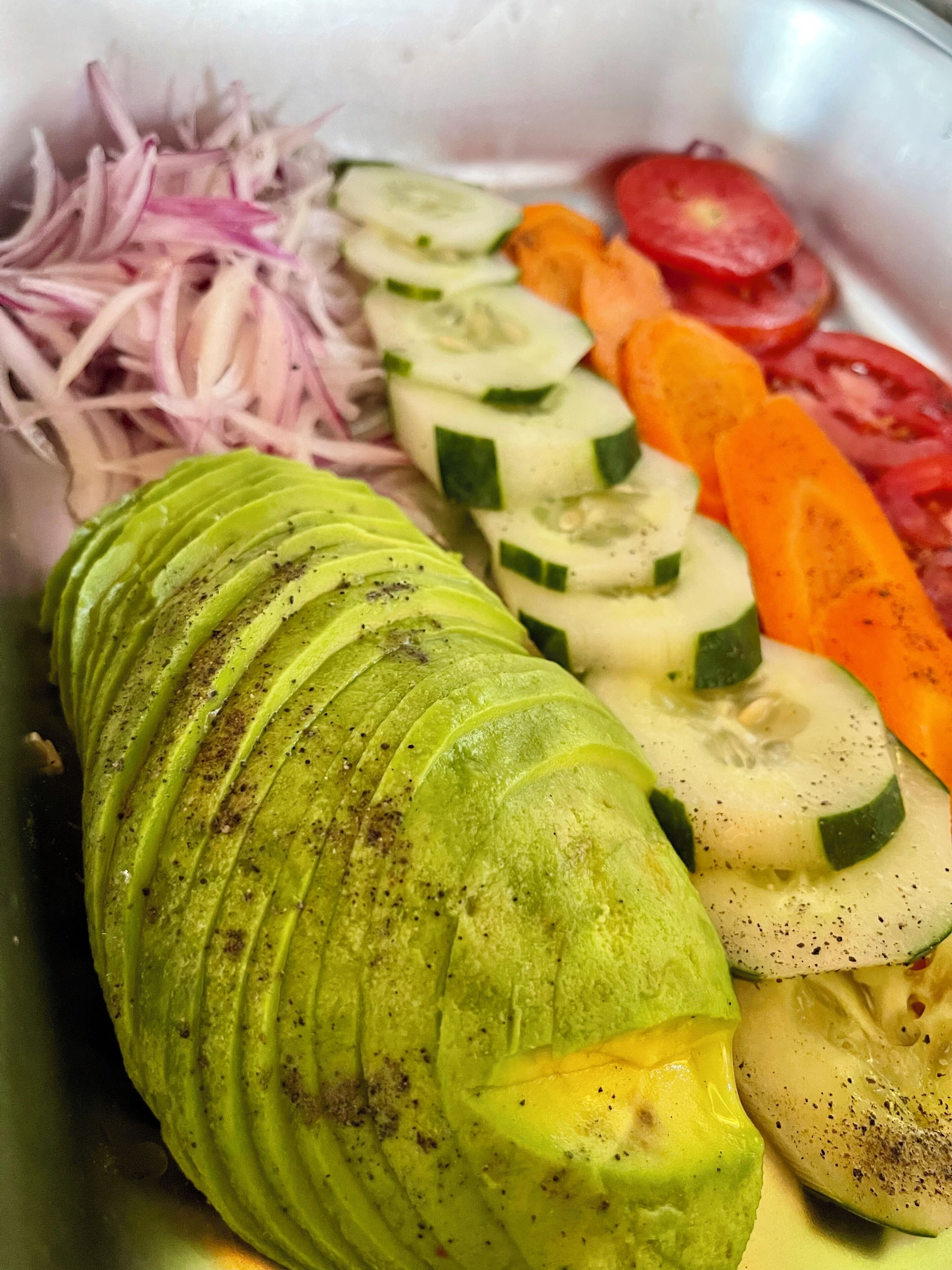 A variety of sliced vegetables including avocado, cucumber, tomato, and carrot in a metal container.