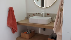 A modern bathroom with a white sink, a round mirror with a light, and a wooden shelf.