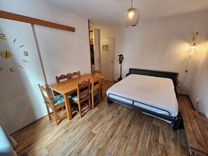 A room with a wooden dining table and chairs, a bed with a metal frame, a wall clock, and a hanging light fixture.