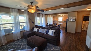 A spacious living room with a dark brown sectional sofa, a ceiling fan, and a view of the kitchen and dining area through large windows. - Lakefront Escape with Hot Tub (Quitman)