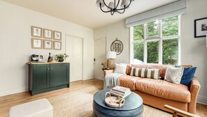 A cozy living room with a leather sofa, a coffee table, and a cabinet. There are framed pictures on the wall and a window with a view of trees. - A beautifully restored Victorian lodge set at the entrance to the picturesque Samlesbury Hall estate (Preston)