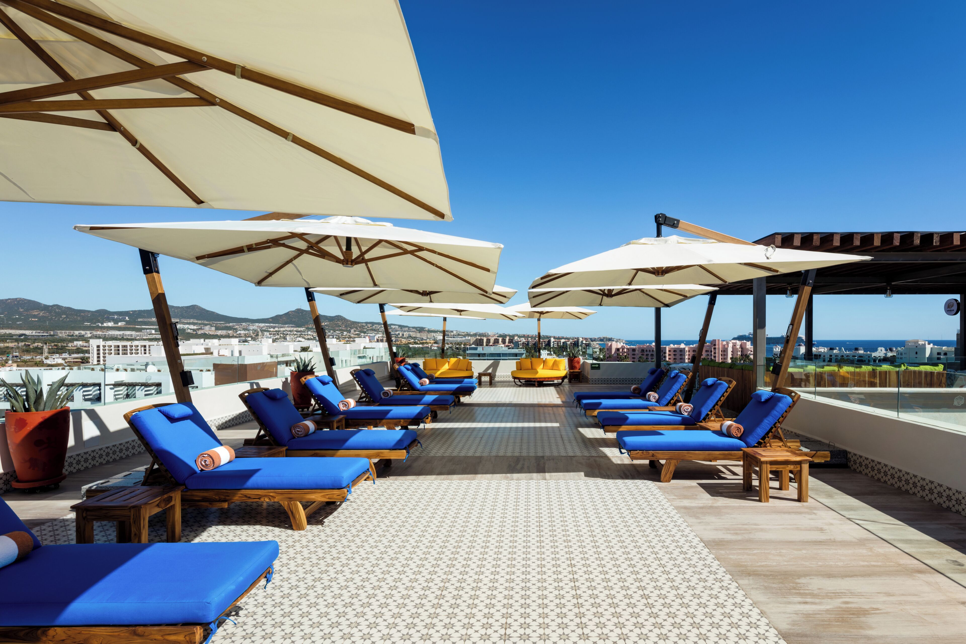 A rooftop lounge area with blue lounge chairs and white umbrellas, offering a view of the city and mountains.