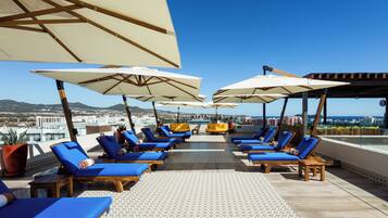 A rooftop lounge area with blue lounge chairs and white umbrellas, offering a view of the city and mountains.