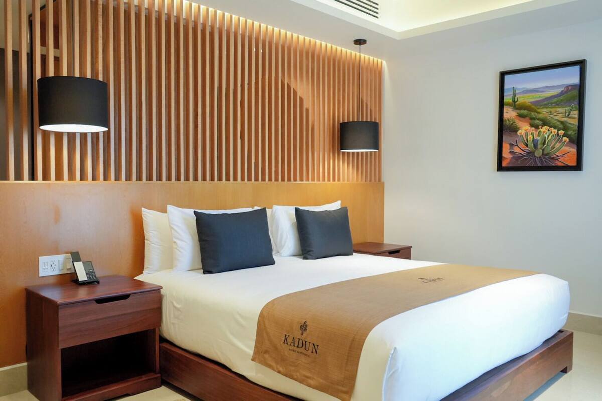 Hotelkamer met een bed, een houten hoofdbord, twee bedlampen, een nachtkastje, een schilderij van een woestijnlandschap en een plaid met de naam van het hotel.