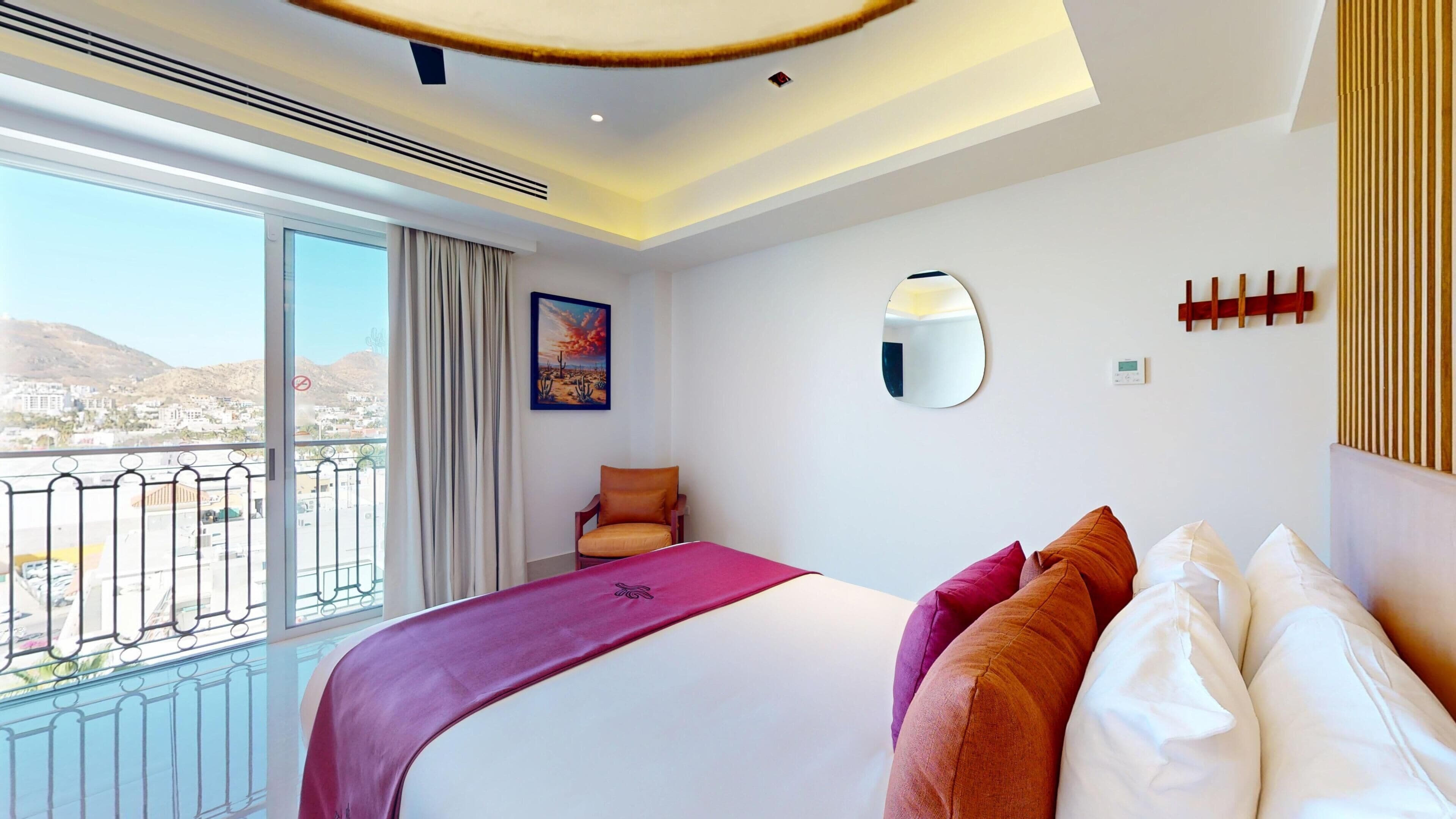 A hotel room with a large bed, a view of the city, a painting, a mirror, and a balcony with a railing.