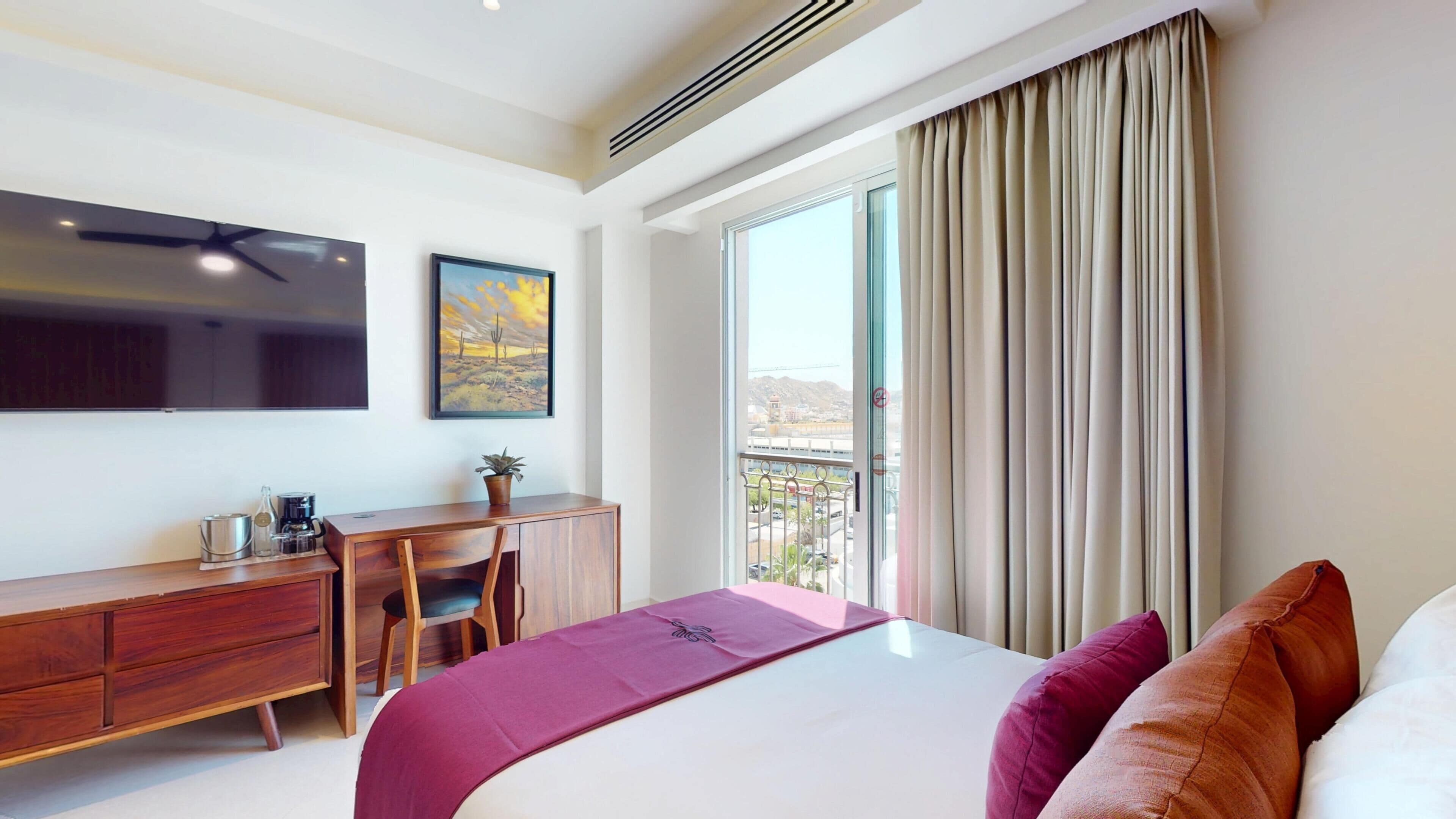 Habitación de hotel con cama, escritorio, silla, televisor y balcón con cortinas.
