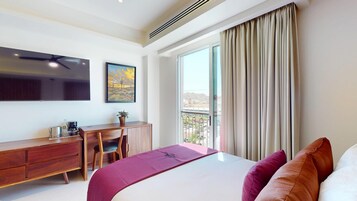 Habitación de hotel con cama, escritorio, silla, televisor y balcón con cortinas.