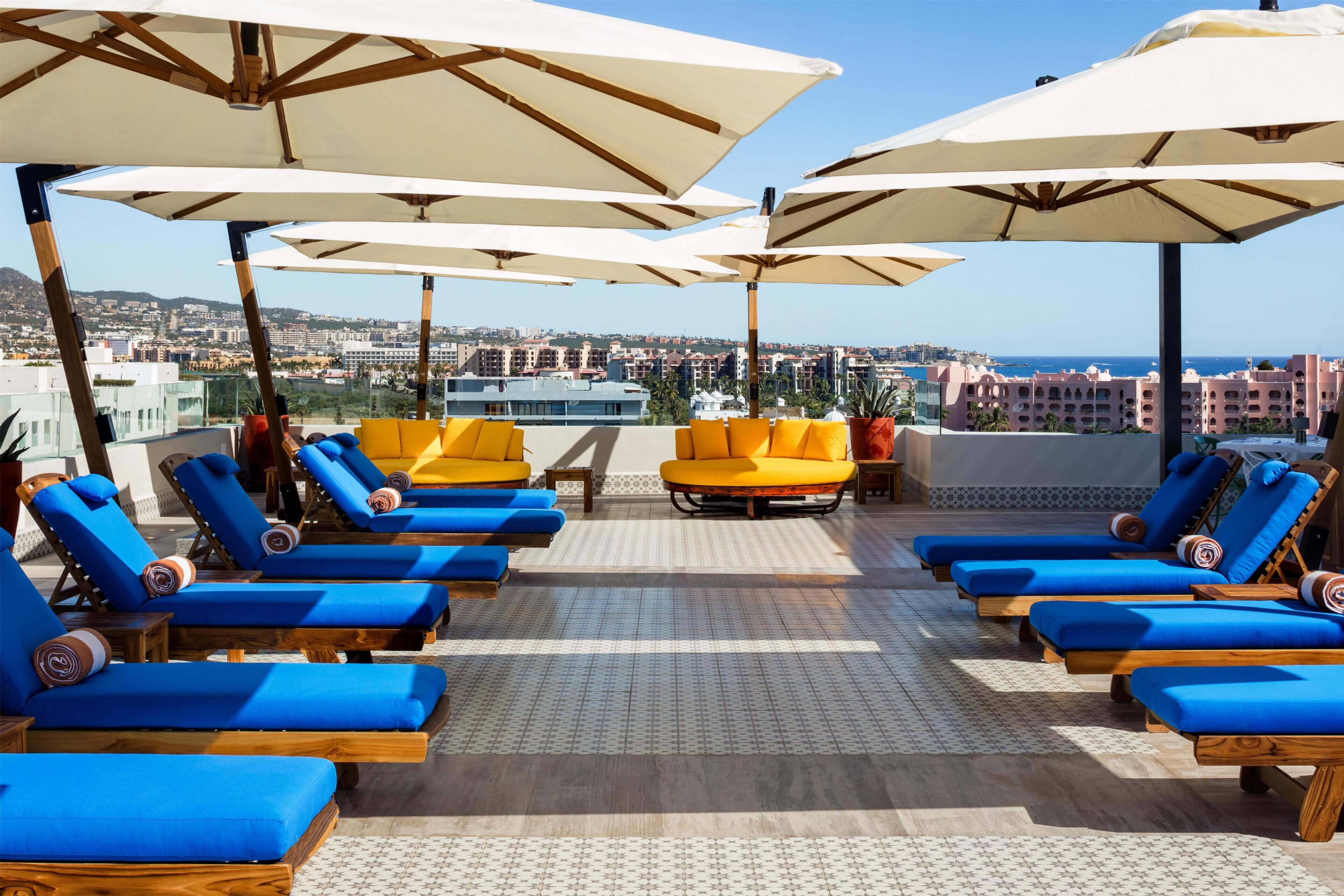 A rooftop lounge area with blue and yellow lounge chairs, white umbrellas, and a city view.