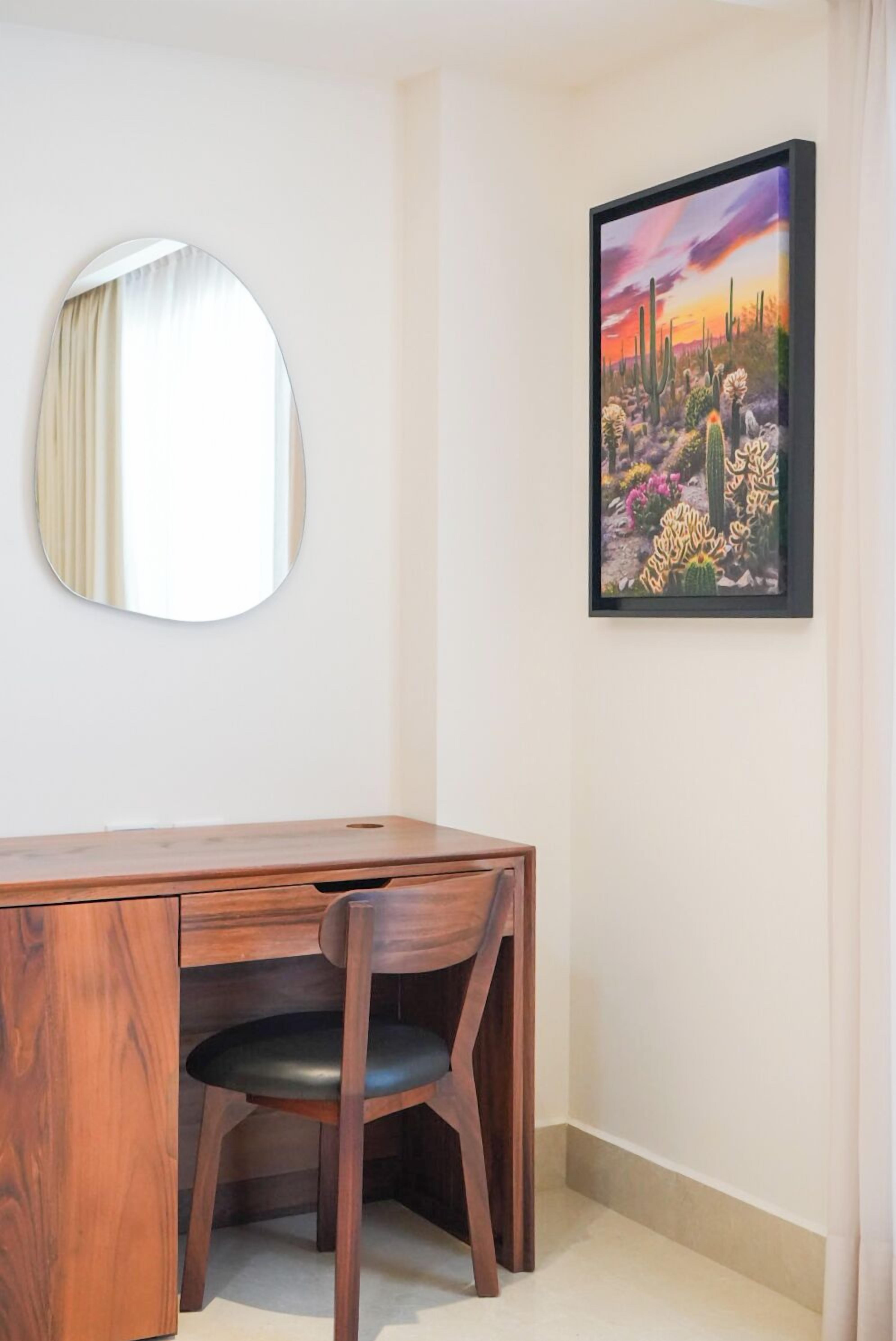 Una habitación con un escritorio de madera, un espejo redondo y un cuadro enmarcado de un paisaje desértico.