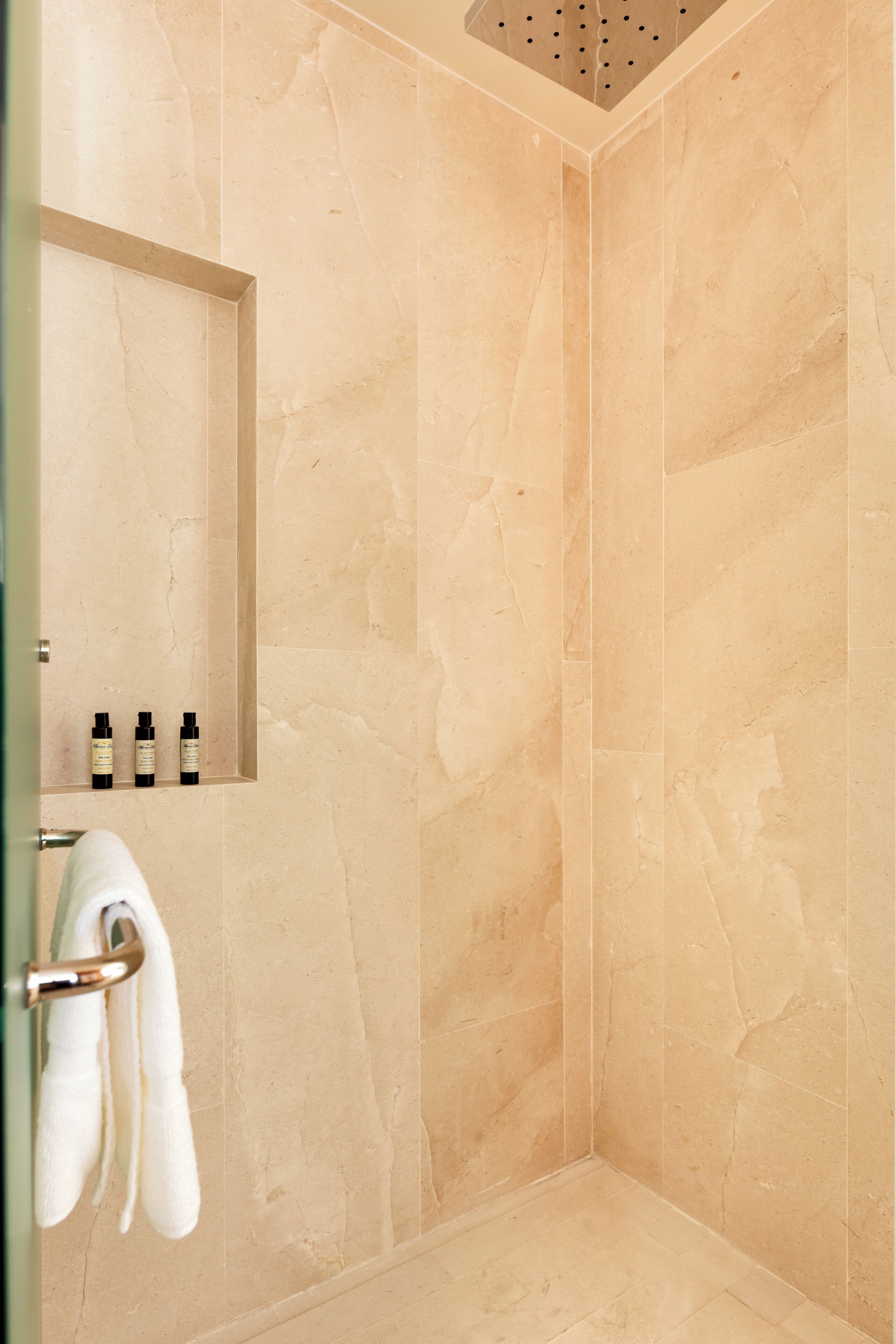 A bathroom with beige marble walls, a shower with a ceiling-mounted rainhead, and a towel rack with a white towel.