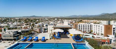 A rooftop pool with blue lounge chairs and umbrellas, overlooking a cityscape with buildings and mountains.