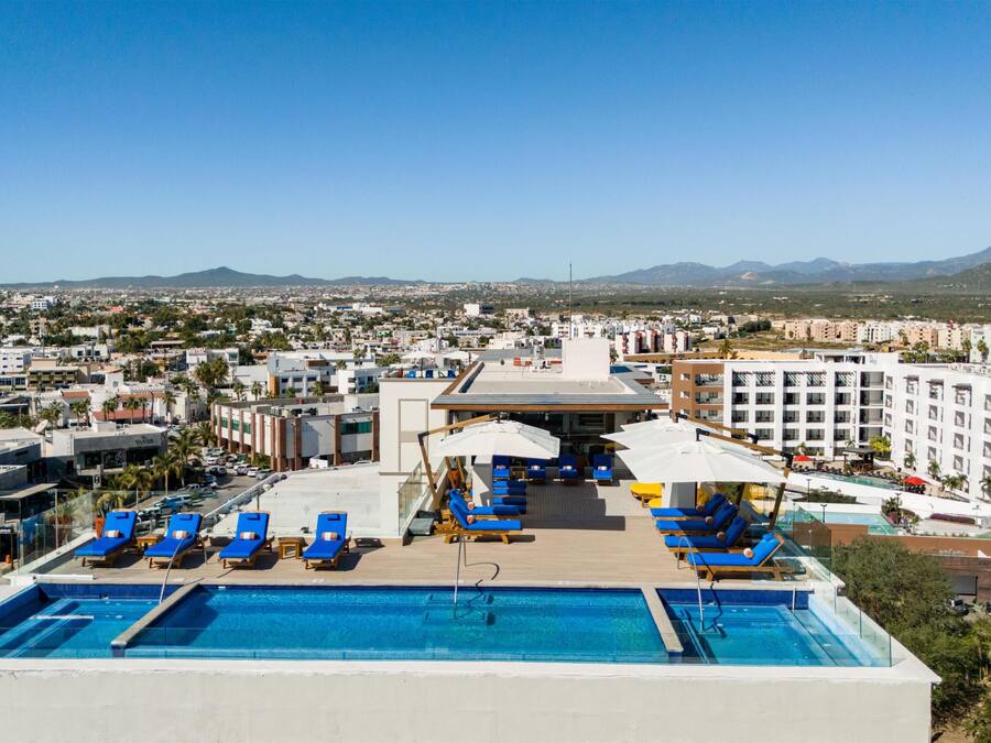 A rooftop pool with blue lounge chairs and umbrellas, overlooking a cityscape with buildings and mountains.