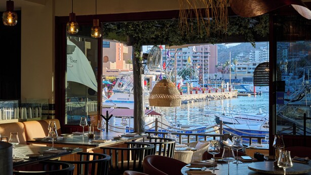 Un restaurante con mesas y sillas, vistas a un puerto deportivo con barcos y un amplio ventanal con vistas al agua.