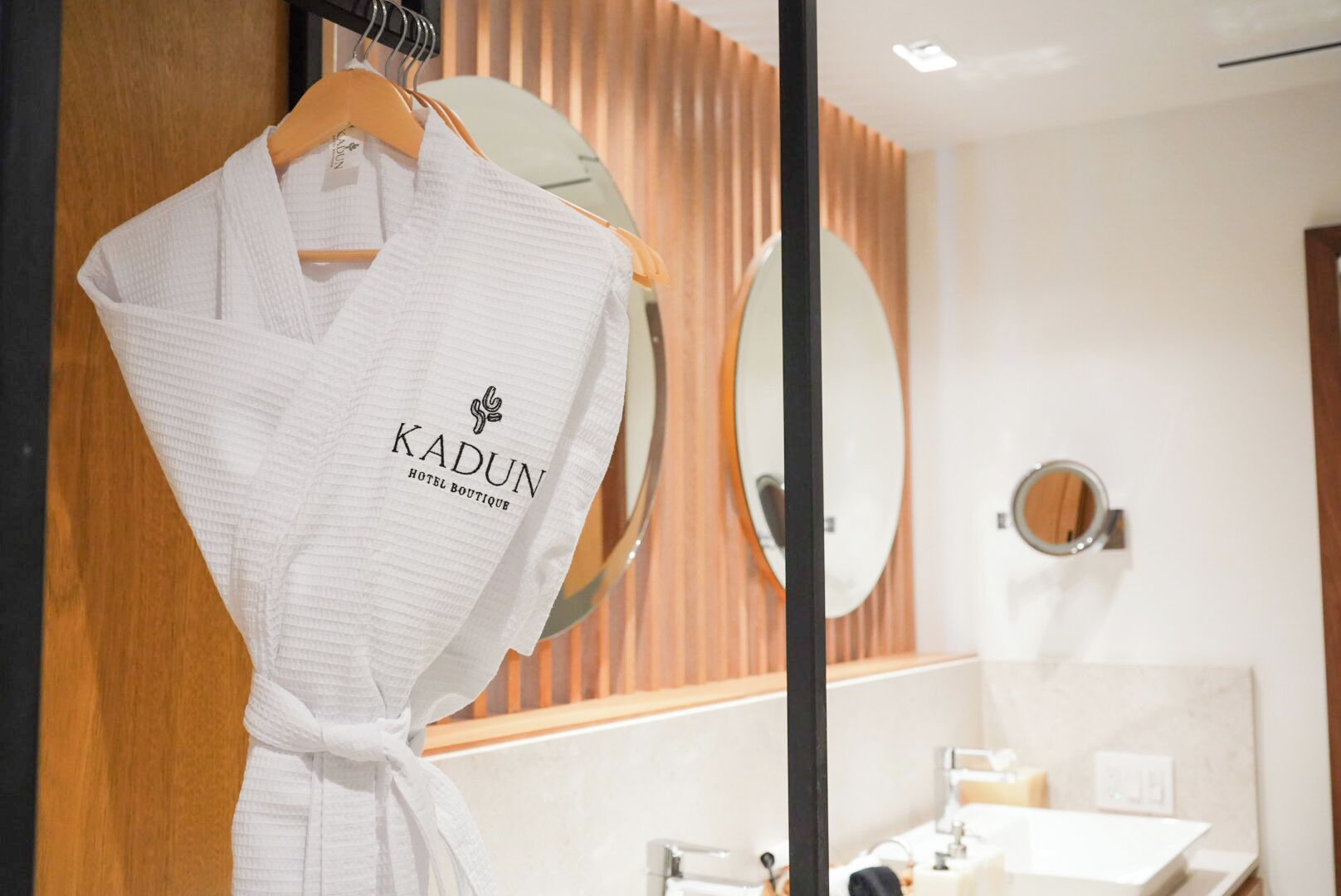 A white robe with "KADUN HOTEL BOUTIQUE" logo hanging on a wooden hanger in a modern bathroom with wooden paneling, a round mirror, and a sink.