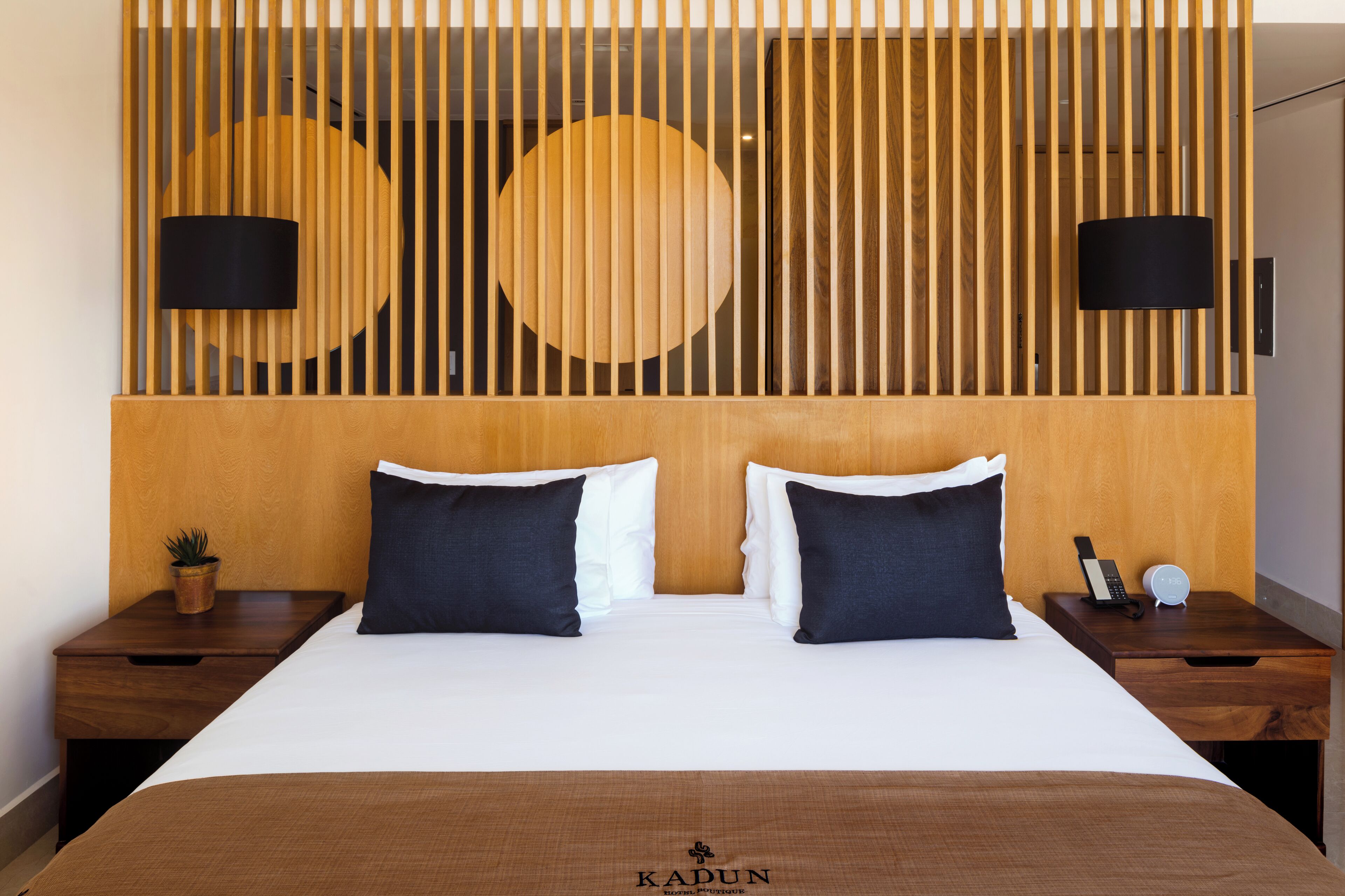 A hotel room with a wooden headboard, two black lamps, a bed with white and dark blue pillows, a small plant on the nightstand, and a logo on the bedspread.