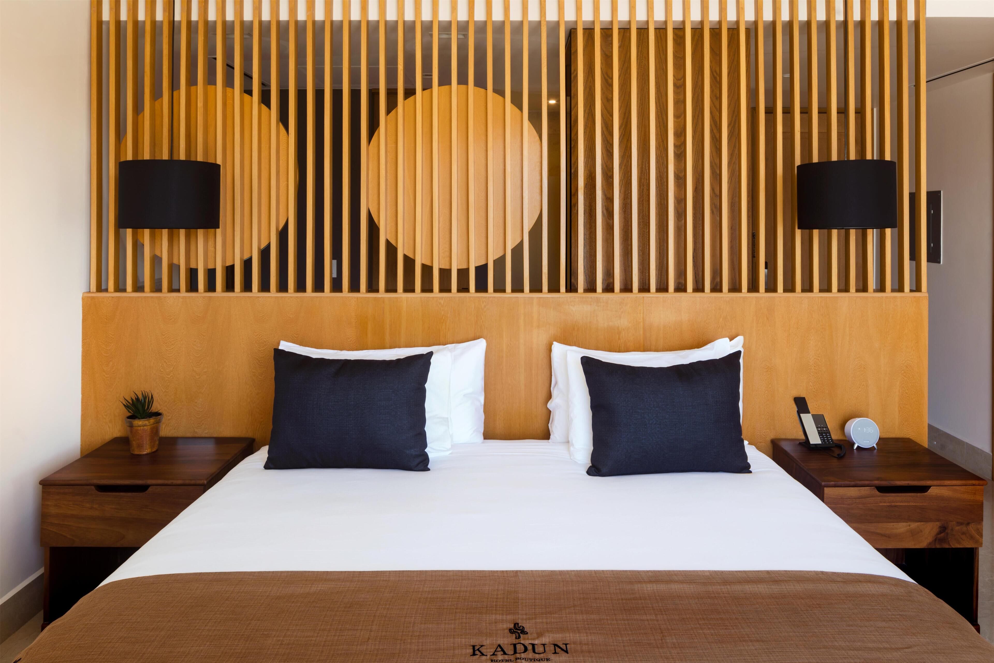 Quarto de hotel com cabeceira de madeira, duas luminárias pretas, cama com almofadas brancas e escuras, mesa de cabeceira com telefone e relógio, e uma pequena planta no criado-mudo.