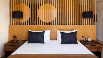 Quarto de hotel com cabeceira de madeira, duas luminárias pretas, cama com almofadas brancas e escuras, mesa de cabeceira com telefone e relógio, e uma pequena planta no criado-mudo.