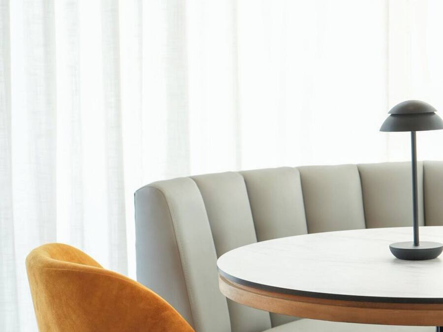 A round table with a black lamp, a mustard-colored chair, and a wicker chair in a room with white curtains.