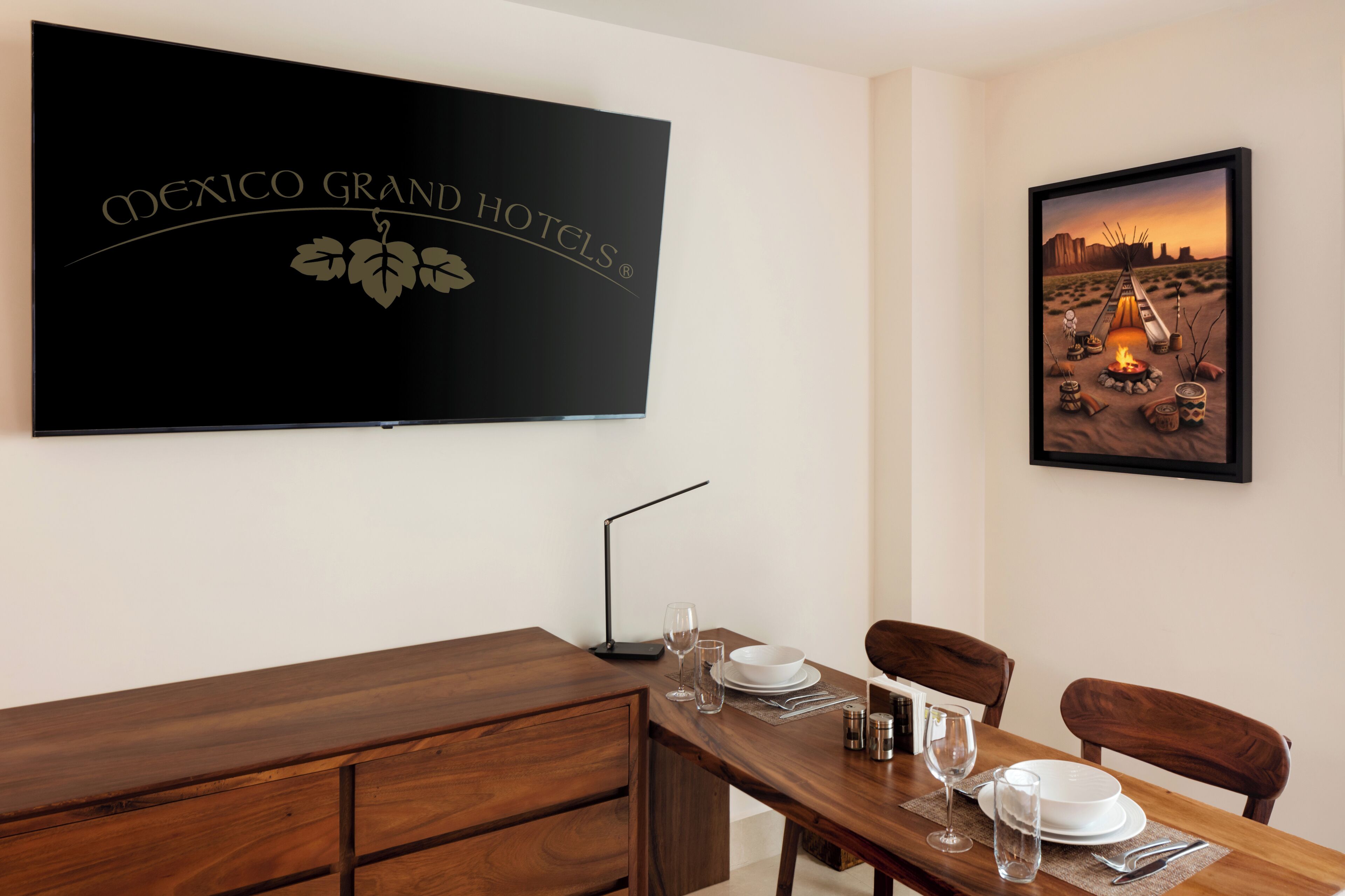 A hotel room with a wooden dining table set for two, a flat-screen TV on the wall, and a framed picture of a desert scene.