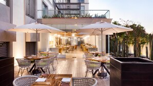 Un espacio al aire libre para comer con sillas y mesas de mimbre, sombrillas y vistas al interior del edificio.
