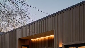 A modern building with a dark brown exterior, a black garage door, and a well-maintained front yard with small trees and a concrete pathway. - The Brown Oak: Nature & Splendor (Conroe)