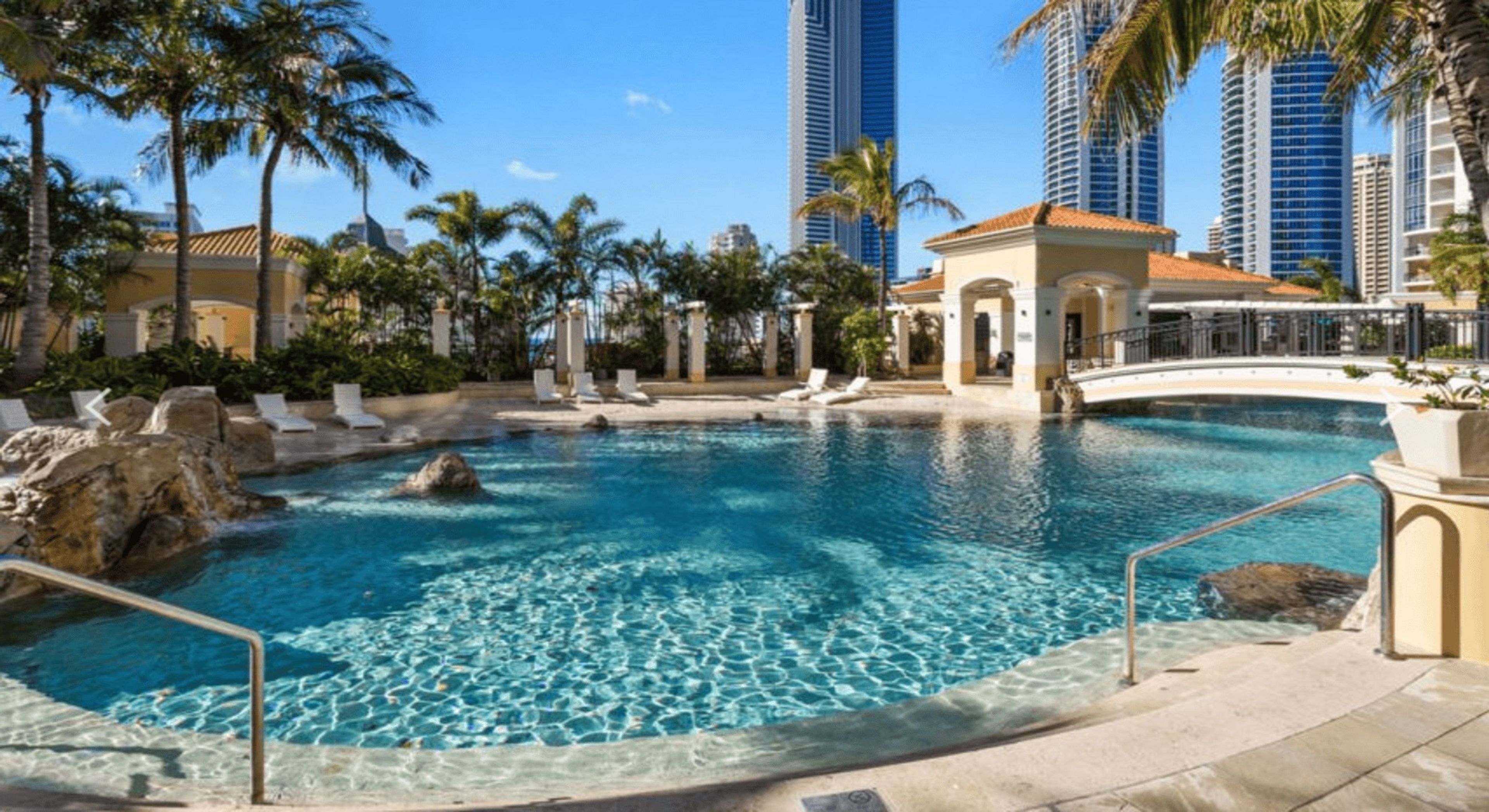 A pool area with clear water, lounge chairs, palm trees, and modern buildings in the background.