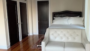 A bedroom with a large bed, a white sofa, wooden flooring, and a door.