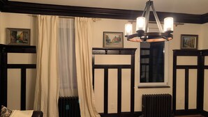 A room with a wooden floor, a black and white paneled wall, a radiator, a window with a curtain, and a chandelier.