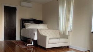 A bedroom with a bed, a white armchair, a wooden floor, and a window with curtains.