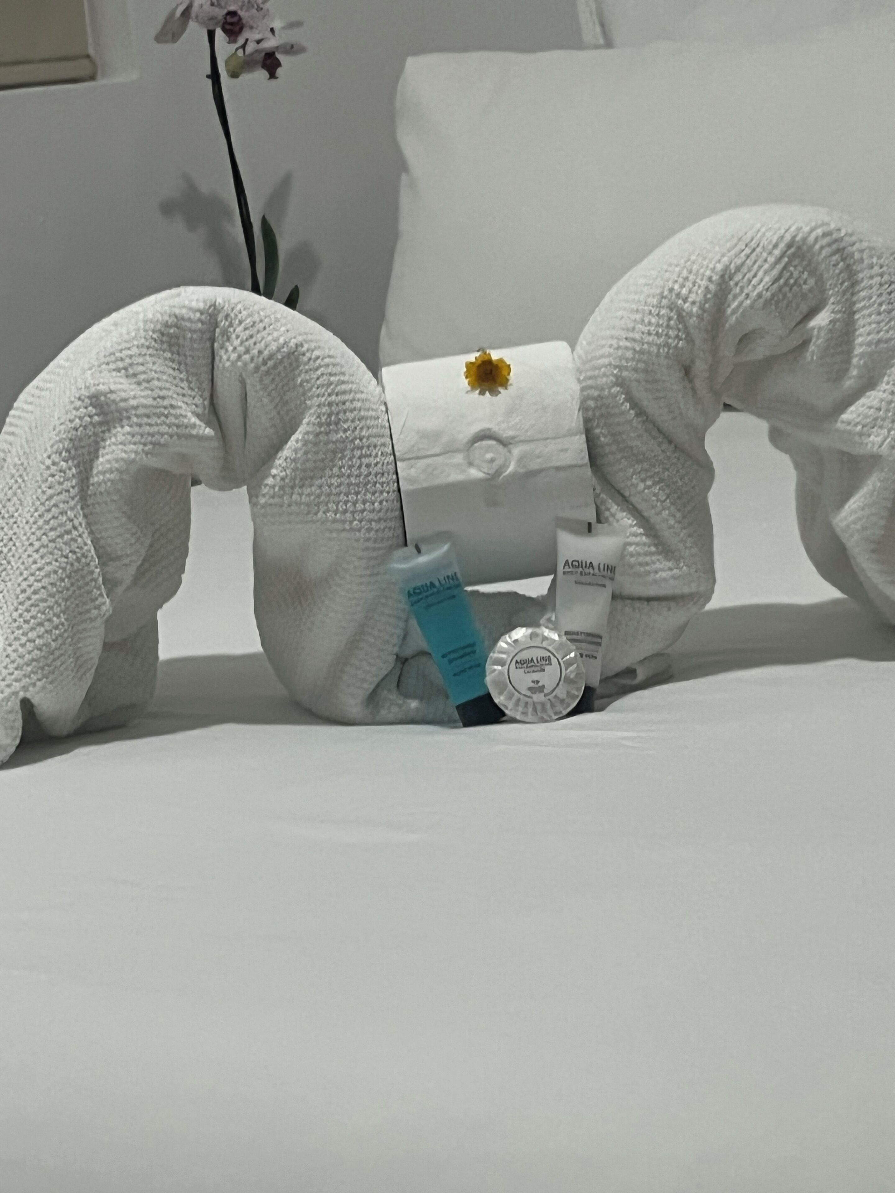 Une sculpture en forme de serviette blanche est posée sur un lit, accompagnée d’un oreiller, d’une petite fleur et d’articles de toilette.