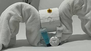 A white towel sculpture on a bed with a pillow, a small flower, and toiletries.