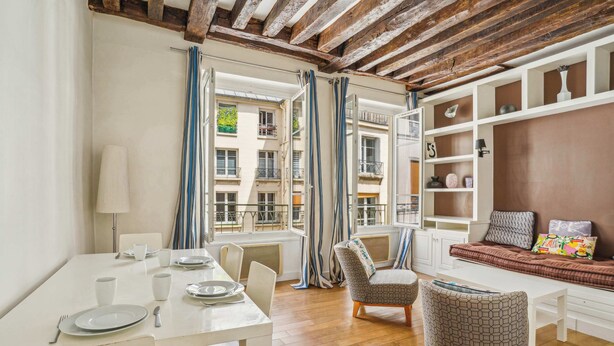 A dining area with a white table set for four, a sofa with cushions, and a bookshelf with decorative items.