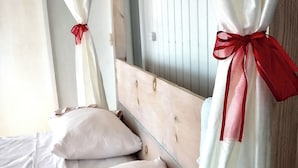 Un lit bien fait, avec une literie blanche et une tête de lit en bois, des rideaux noués avec des rubans rouges, et un mur en bois à l’arrière.