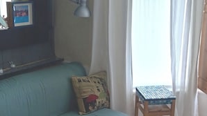 Une pièce avec un canapé bleu, un petit tabouret en bois, une lampe et une fenêtre avec des rideaux.