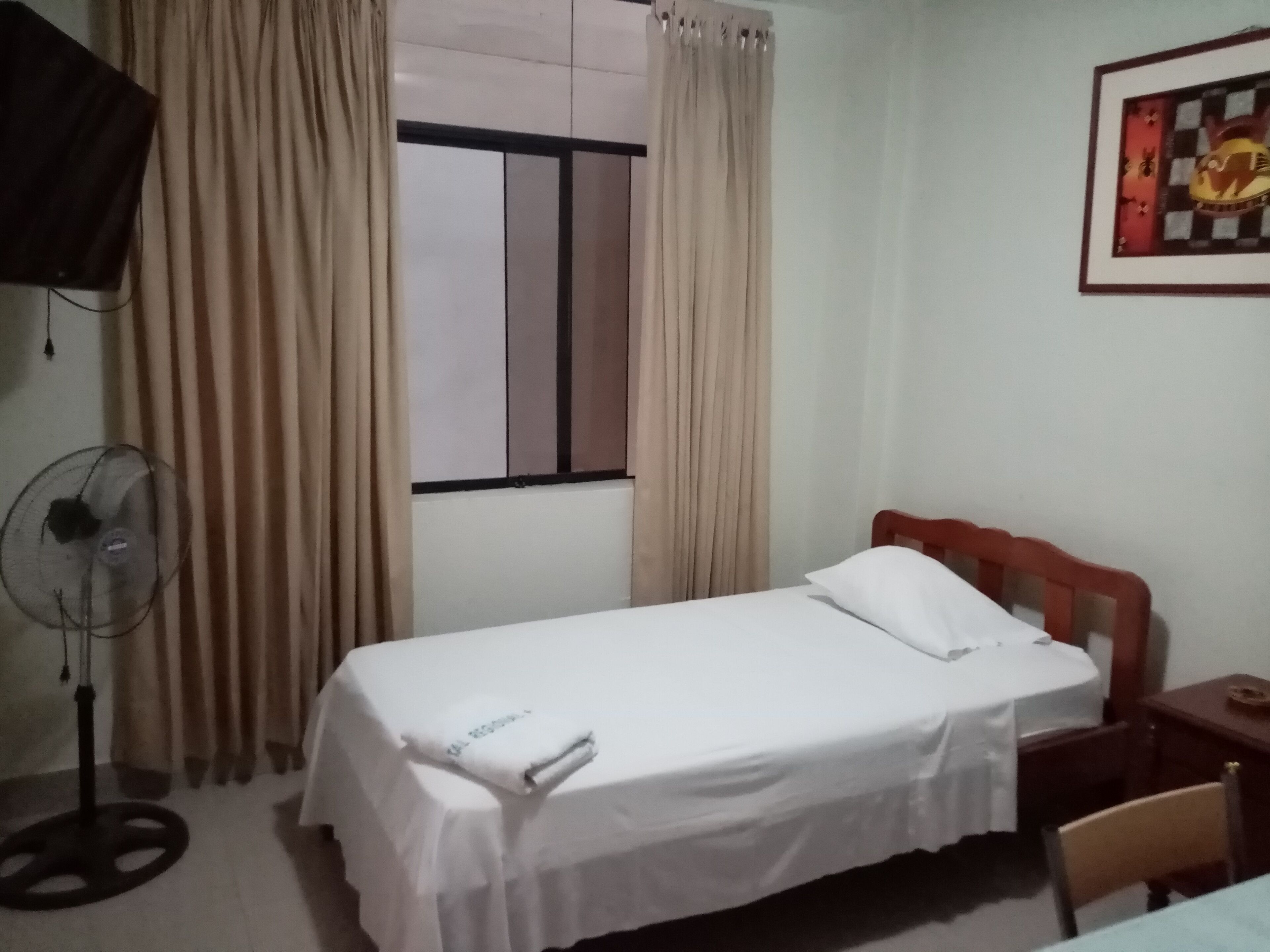 A hotel room with a single bed, a bedside table, a chair, a standing fan, and a window with curtains.