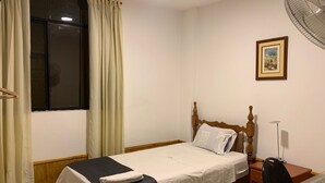 A single bed with a wooden headboard, a bedside table, a chair, a desk, a standing fan, a window with curtains, and a framed picture on the wall. - hostal regional (Piura)