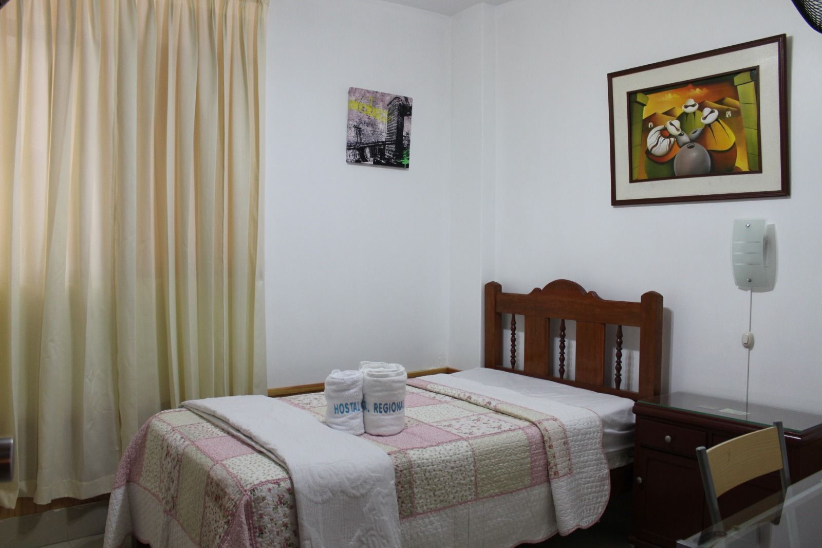 A hotel room with a bed, a wooden headboard, a painting, a window with curtains, and a bedside table.