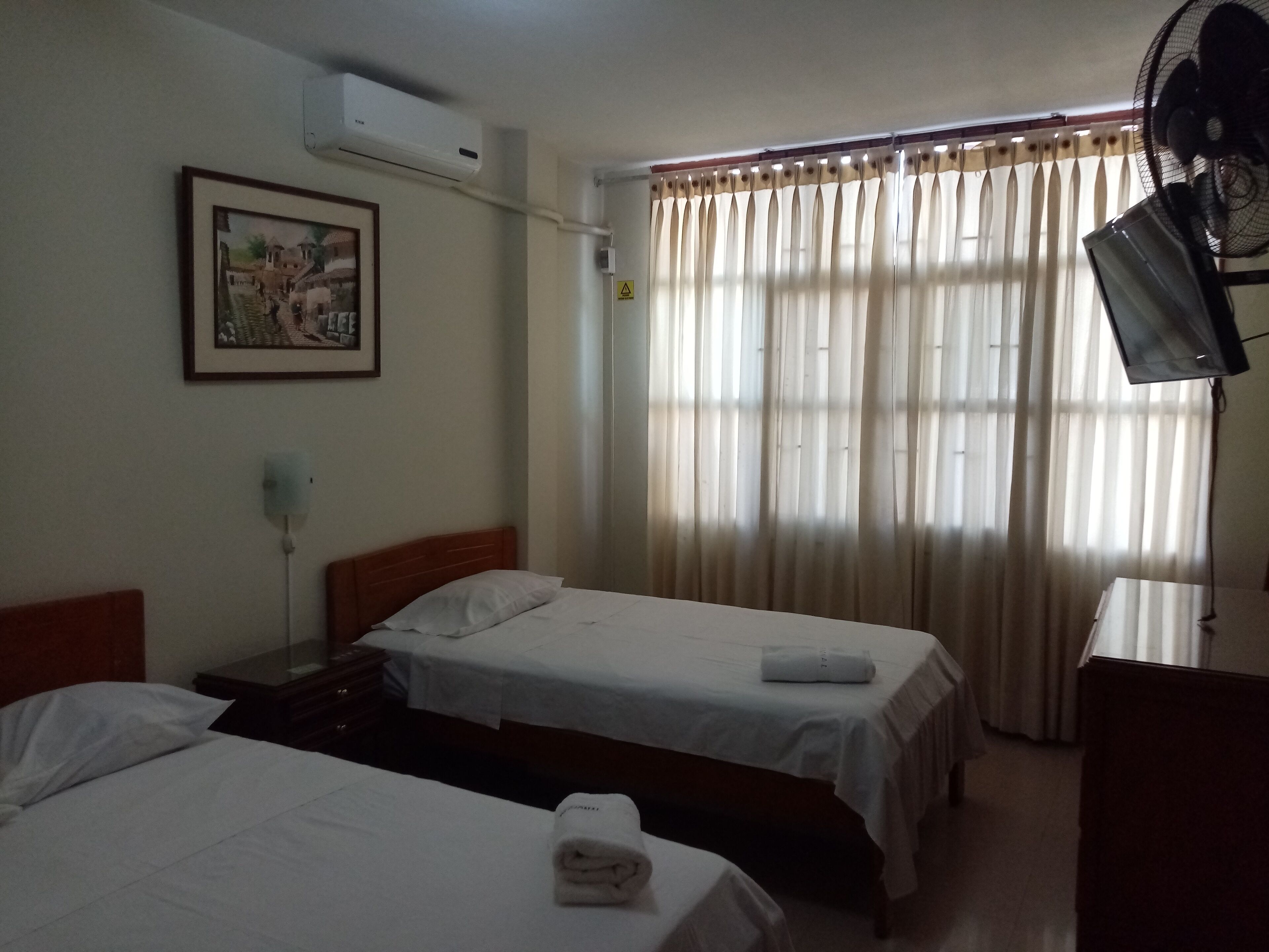 Une chambre d’hôtel avec deux lits simples, une table de chevet avec une lampe, un climatiseur fixé au mur, un tableau encadré et une fenêtre avec des rideaux légers.