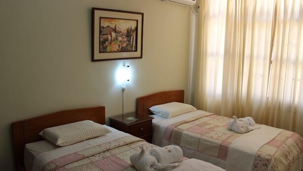 A hotel room with two beds, a nightstand, a wall-mounted lamp, a painting, and a window with curtains. - hostal regional (Piura)