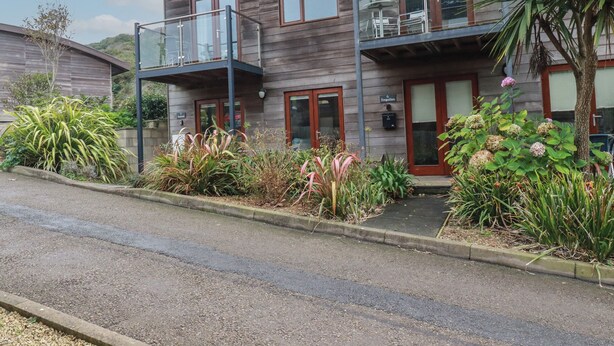 A two-story building with a balcony on the upper floor, a small garden area with various plants, and a wooden door. - Tregullan (Truro)