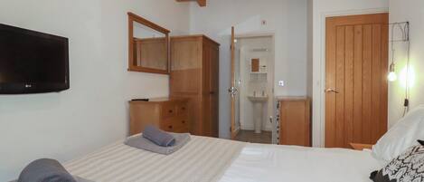 A bedroom with a bed, a TV, wooden furniture, a bathroom visible through an open door, and a light fixture.