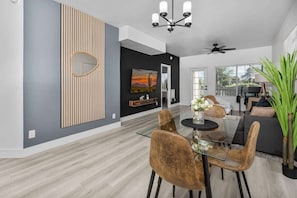 A modern living room with a dining area, featuring a glass table, brown chairs, a ceiling fan, and a large window with a view.