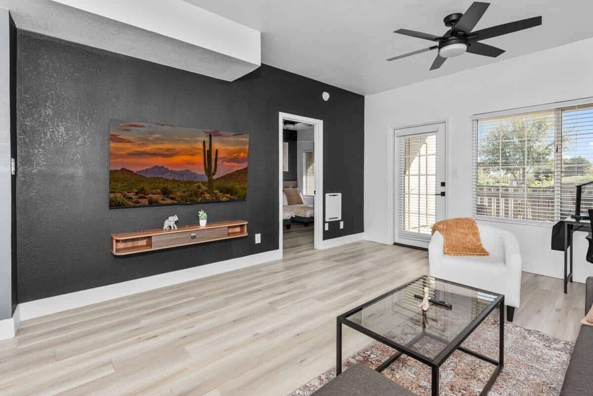 A modern living room with a dark wall, a wooden floor, a glass coffee table, a white armchair, and a ceiling fan.