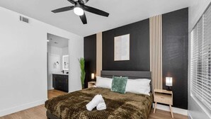 A modern bedroom with a large bed, a ceiling fan, a nightstand with a lamp, and a window with blinds. - Snug 2BR-Pool, BBQ,Prkng, Yoga Rm, Gym, Cabanas (Scottsdale)