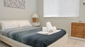 Una cama bien hecha con ropa de cama gris y blanca, una mesita de noche de madera con una lámpara, y un aparador de madera con un objeto pequeño encima.