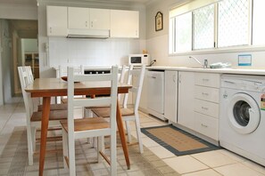 Une cuisine compacte avec des meubles blancs, un four à micro-ondes, un lave-vaisselle et un lave-linge. On y trouve une table à manger avec des chaises et une fenêtre qui laisse entrer la lumière naturelle.