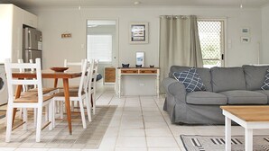 Una sala de estar moderna con un sofá gris, una mesa de comedor de madera y sillas. Hay una zona de cocina con un refrigerador y una mesa de comedor.