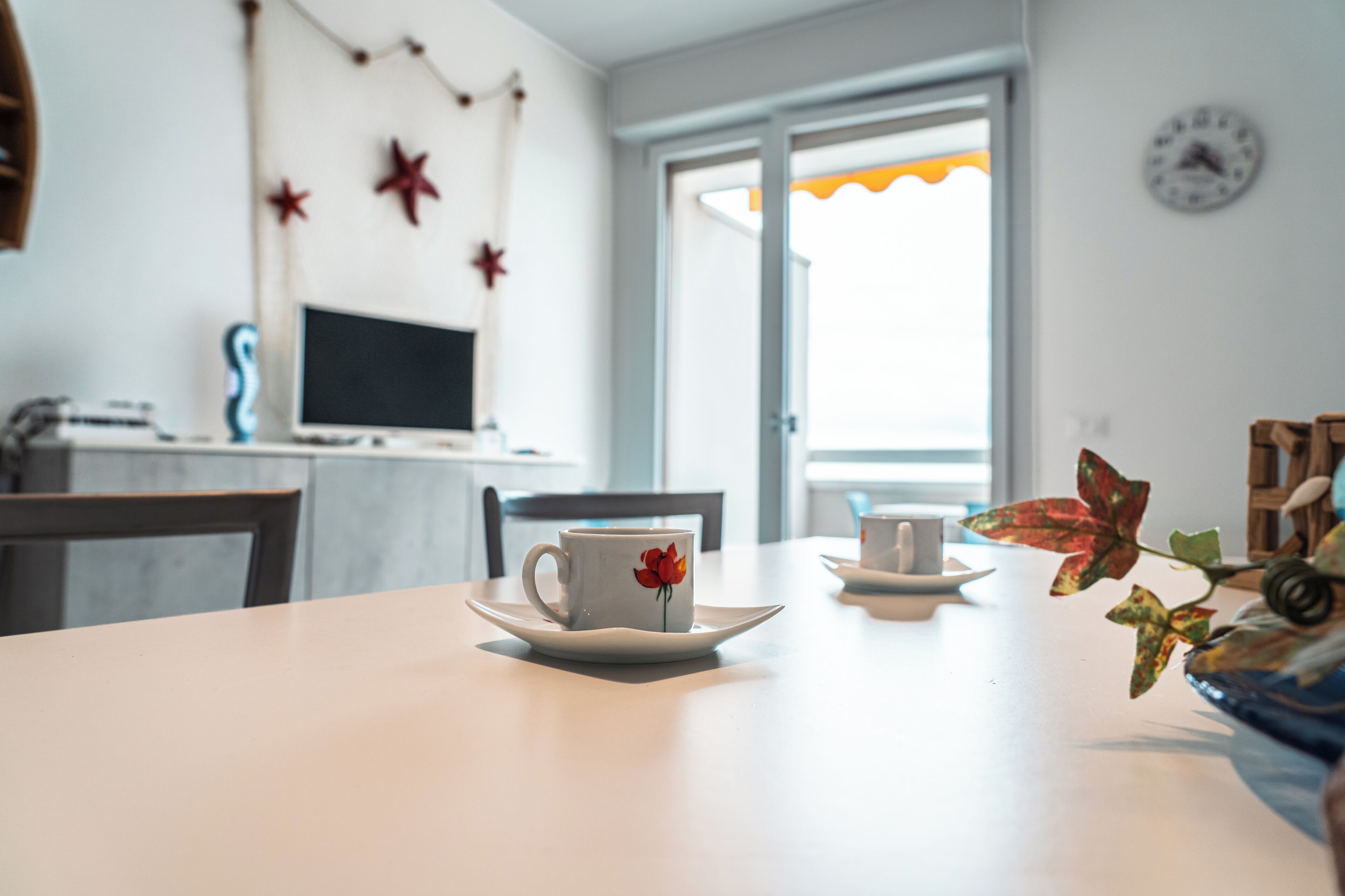 A dining table with a white plate and a cup with a red flower design. There is a window with a view of the outside, a clock on the wall, and a plant with red leaves.