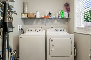 A laundry room with two front-loading washers, a shelf above containing cleaning supplies, and a window with blinds.