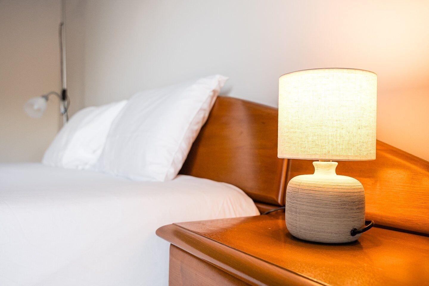 Un lit avec une literie blanche et une tête de lit en bois, une table de chevet avec une lampe à texture intéressante et un luminaire fixé au mur.